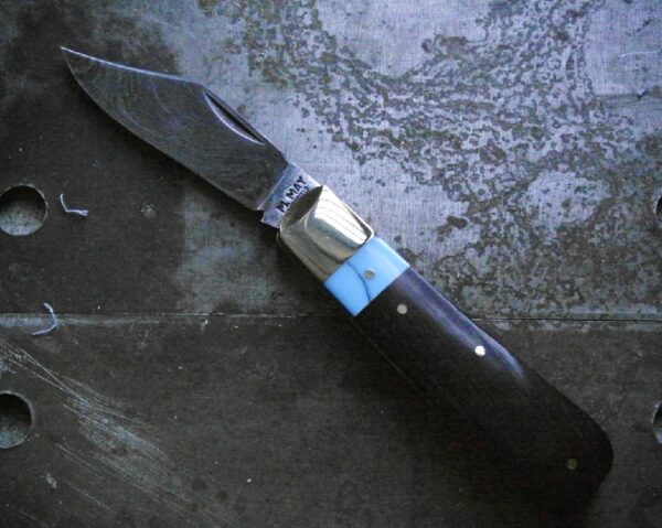 A handmade pocket knife featuring a damascus steel blade with swirled patter, paired with a handle made of ebony wood and a turquoise collar. The bolster is polished brass, giving the knife a striking and elegant appearance.